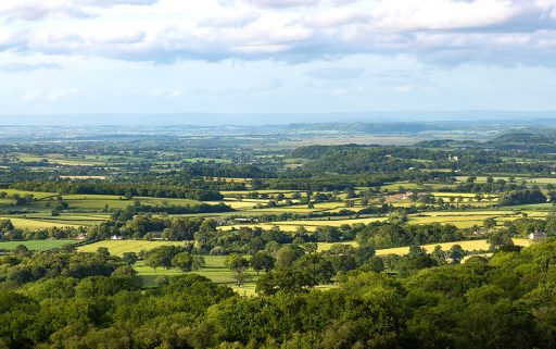 A wide landscape with a patchwork of fields and hedgerows and hills in the background