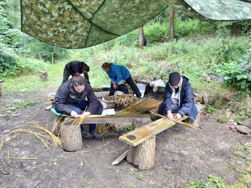Somerset Nature Connections nature and wellbeing course, 4 people doing weaving on log benches under a tarpaulin