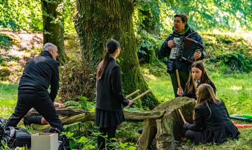 An accordion player with children playing percussion on tree stumps in the woods